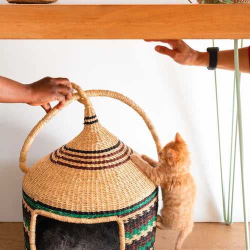 A natural woven basket cat tree with a pointed lid and arched handles, shown in a home setting with a ginger cat climbing on it.